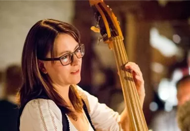 Eine Frau spielt auf einem Kontrabass in einer stimmungsvollen Umgebung. Sie trägt eine Brille und wirkt konzentriert.