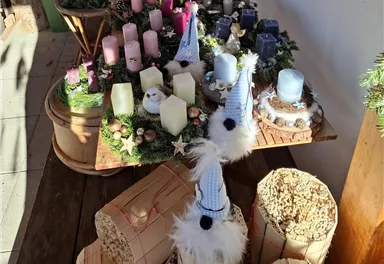 Eine dekorative Anordnung von Kerzen in verschiedenen Farben, umgeben von Tannenzweigen und Weihnachtsornamenten. Im Vordergrund sind festliche Figuren mit Mützen und flauschigen Bärten zu sehen.