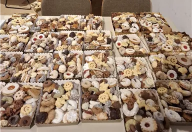 Eine große Anzahl von Keksen in verschiedenen Formen und Farben, gleichmäßig auf mehreren Platten angeordnet. Die Platten liegen auf einem Tisch und zeigen eine Vielzahl von süßen Leckereien.