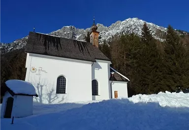 A small white chapel stands in the snow, surrounded by tall mountains and coniferous trees. The clear blue sky provides a beautiful contrast to the wintry landscape.