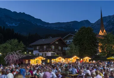 Ein lebhaftes Fest bei Nacht mit vielen Menschen und festlich beleuchteten Ständen. Im Hintergrund erheben sich Berge und eine Kirche mit beleuchtetem Turm.