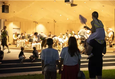 Eine Familie schaut sich ein Konzert auf einer Bühne an. Im Hintergrund spielt eine Band, während die Zuschauer entspannt den Abend genießen.