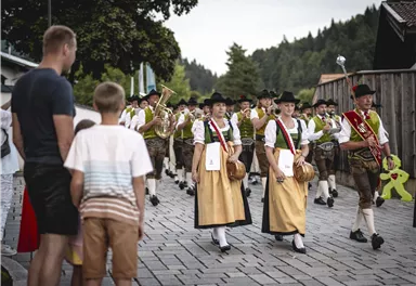 Eine traditionelle Umzug mit Musikkapelle und Menschen in Trachten. Im Hintergrund stehen Zuschauer und genießen das Fest.