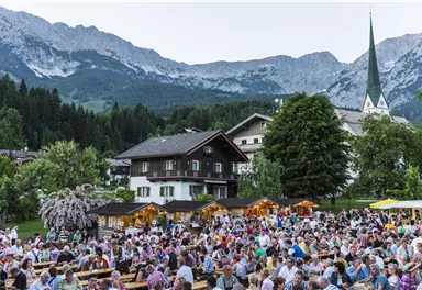 Eine große Menschenmenge sitzt in einem malerischen Dorf unter den Bergen. Umgeben von Bäumen und traditionellen Gebäuden, genießen die Besucher eine festliche Atmosphäre.