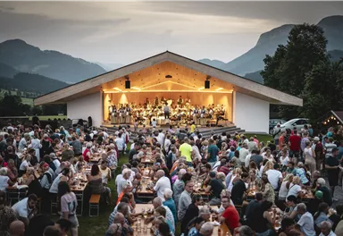 Eine große Menschenmenge genießt ein Konzert im Freien. Auf der Bühne spielt eine Musikgruppe, während im Vordergrund viele Tische und Stühle stehen.