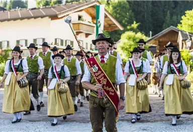 Eine traditionelle Festparade mit Menschen in Trachten. Im Vordergrund führt ein Mann mit einem Zepter die Gruppe an.