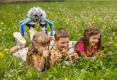 Vier Kinder liegen auf einer Wiese und schauen mit Lupe im Gras. Ein buntes, kreativ gestaltetes Wesen beobachtet sie von hinten.