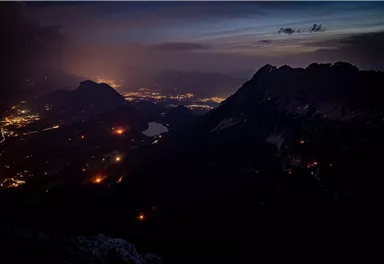 An impressive mountain landscape at night with shimmering lights of the city in the valley. The sky is streaked with soft clouds and shows a hint of twilight.