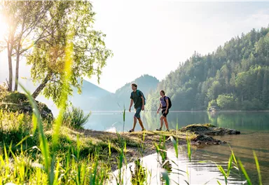 Zwei Wanderer spazieren am Ufer eines klaren Sees. Umgeben von grünen Bäumen und Hügeln im Hintergrund.