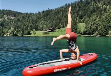 Eine Person macht einen Handstand auf einem Stand-Up-Paddleboard im klaren Wasser eines Sees. Im Hintergrund sind grüne Bäume und hügelige Landschaften zu sehen.