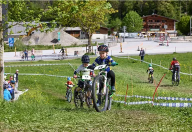 Kinder fahren mit Mountainbikes einen Wettkampf auf einer grünen Wiese. Im Hintergrund sind Zuschauer und Bäume sichtbar.