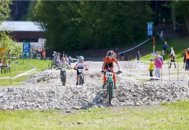 Zwei Fahrradfahrer fahren über einen technischen Parcours mit Steinen. Im Hintergrund sind Zuschauer und ein grünes Umfeld zu sehen.