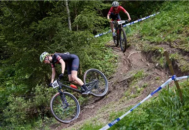 Zwei Mountainbiker fahren einen schmalen, kurvigen Weg durch einen grünen Wald. Die Strecke ist mit Absperrungen markiert und zeigt eine dynamische Wettkampfsituation.