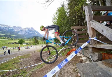 Ein Radfahrer springt mit seinem Mountainbike über ein Hindernis auf einem Trainingsgelände. Im Hintergrund sind eine grüne Landschaft und Berge zu sehen.