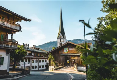 Eine malerische Dorfstraße mit traditioneller Architektur und einer hohen Kirchturmspitze. Im Hintergrund sind sanfte Berge und ein klarer Himmel zu sehen.