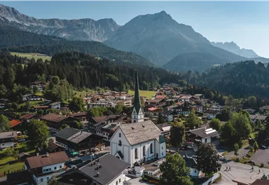 A picturesque village view with a white church in the foreground and the Alps in the background. The surroundings are green and mountainous, ideal for nature lovers.