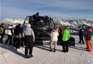 A group of people is standing around a snow groomer in the mountains. In the background, snow-covered peaks and a clear sky can be seen.