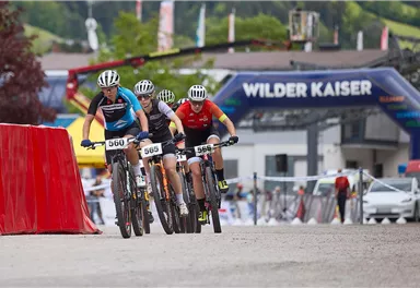 Ein spannendes Mountainbike-Rennen mit mehreren Radfahrern, die um die Wette fahren. Im Hintergrund sind festliche Banner und eine grüne Landschaft zu sehen.