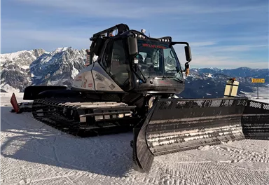 A snow groomer stands on a snowy mountain with impressive mountain peaks in the background. The sky is clear and blue.