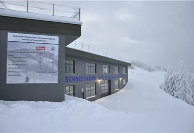 Ein modernes Gebäude in einer verschneiten Landschaft. Schilder mit technischen Informationen zur Schneeproduktion sind sichtbar.
