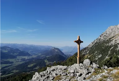 Ein Holzkreuz auf einem Berggipfel mit majestätischem Ausblick auf das Tal. Der Himmel ist klar und blau, und die Berge sind im Hintergrund sichtbar.