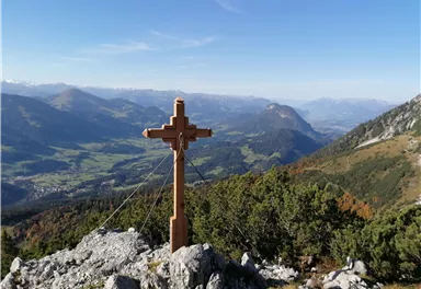 Ein Holzkreuz steht auf einem Felsen mit Blick auf ein schönes Tal. Die Berge und der klare Himmel im Hintergrund schaffen eine beeindruckende Aussicht.