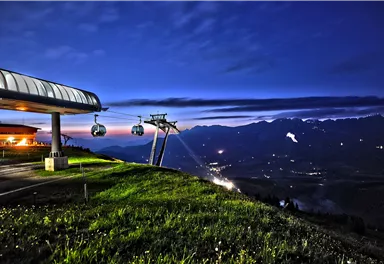 Eine Bergbahn mit Gondeln bei Nacht in der Natur. Im Hintergrund sind die silhouettierenden Berge und der klare Nachthimmel zu sehen.