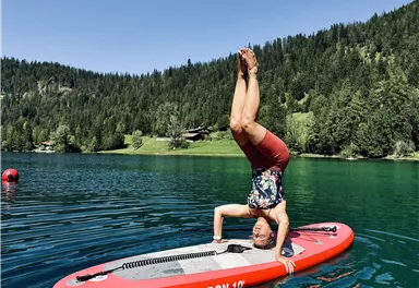 Ein Mann macht einen Handstand auf einem roten Stand-up-Paddleboard. Im Hintergrund sind grüne Wälder und ein klarer, blauer See zu sehen.