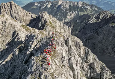 Eine Gruppe von Wanderern erklimmt eine felsige Berglandschaft. Im Hintergrund sind majestätische Berge und ein klarer Himmel zu sehen.