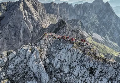 Ein atemberaubender Blick auf einen zerklüfteten Gebirgskamm mit vielen Menschen auf dem Gipfel. Die Landschaft ist von majestätischen Bergen und einer klaren Himmelspanne umgeben.