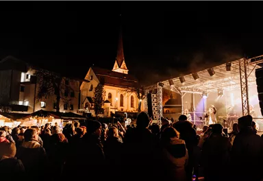 Ein Abendkonzert in einer festlich beleuchteten Stadt. Viele Menschen stehen vor der Bühne und genießen die Musik.