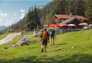 Zwei Wanderer gehen auf einem Weg zu einer Berghütte mit roten Sonnenschirmen. Die Umgebung ist grün und von Bäumen umgeben.