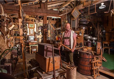 A man stands in an old craft room full of tools and memorabilia. The atmosphere is rustic and traditional.