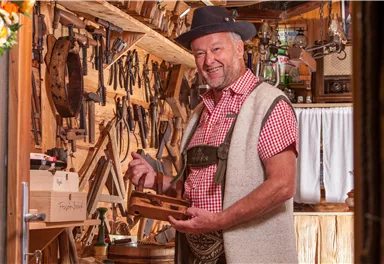 A craftsman in traditional clothing stands in his workshop. Behind him, numerous tools made of wood and metal are visible.