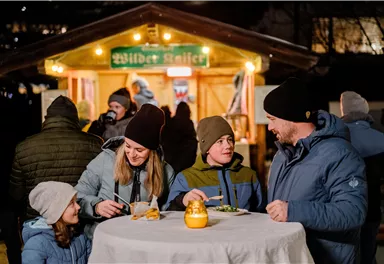 Eine Gruppe von vier Personen steht an einem runden Tisch und genießt Essen und Trinken. Im Hintergrund ist eine beleuchtete Verkaufsbude zu sehen.
