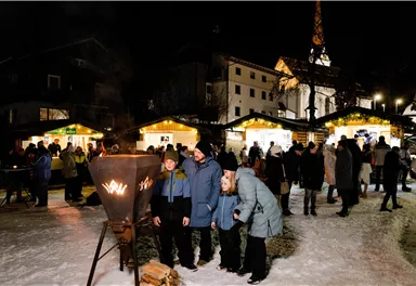 Ein winterlicher Marktplatz bei Nacht mit festlicher Beleuchtung. Menschen genießen die Atmosphäre und die Stände im Hintergrund.