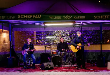 Eine Band spielt live unter einem Zelt in der Nacht. Die Atmosphäre ist festlich mit bunten Lichtern und einer schneebedeckten Umgebung.