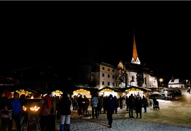 Ein festlicher Weihnachtsmarkt bei Nacht mit vielen Besuchern und stimmungsvollem Licht. Im Hintergrund ist eine Kirche mit einem hohen, spitzen Turm zu sehen.