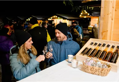 Ein Paar stößt mit Gläsern an und genießt die festliche Atmosphäre eines Marktes bei Nacht. Im Hintergrund sind weitere Menschen und stimmungsvolle Lichter zu sehen.