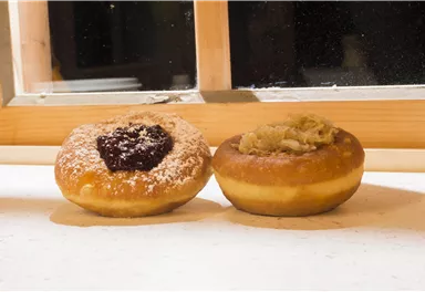 Zwei leckere Donuts auf einem Tisch. Der linke Donut hat eine Füllung aus Marmelade und Puderzucker, der rechte ist mit einer cremigen Füllung.
