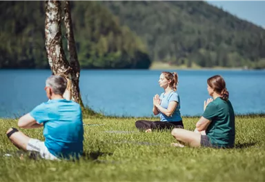 Three people are meditating by the shore of a lake. The surroundings are green and surrounded by trees.