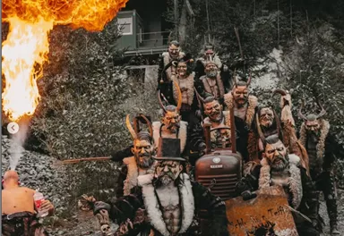 A group of masked people in eerie costumes stands outdoors. One of the participants breathes fire while the others take grim poses.