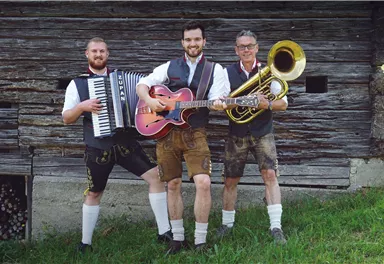 Drei Musiker in traditioneller bayerischer Tracht stehen vor einer Holzwand. Sie spielen ein Akkordeon, eine Gitarre und eine Tuba.