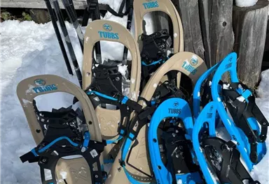Eine Ansammlung von Schneeschuhen in verschiedenen Farben liegt im Schnee. Im Hintergrund sind einige Skistöcke und ein Holzpfosten zu sehen.