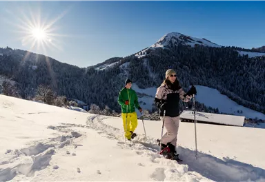 Two people are hiking through a snow-covered landscape in the mountains. The sun is shining brightly in the clear sky.