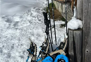 Schneeschuhe und Wanderstöcke liegen im Schnee vor einer Holzscheune. Die Umgebung ist schneebedeckt und die Sonne scheint.