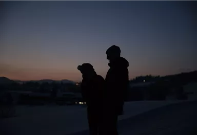 Silhouettes of two people stand in the twilight before a snowy landscape. The sky shows soft colors of the evening dusk.