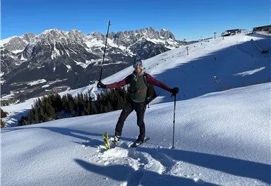 Ein Skifahrer steht im Schnee und posiert mit einem Skistock. Im Hintergrund sind schneebedeckte Berge und ein blauer Himmel zu sehen.