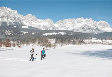 Two people are hiking through a snow-covered landscape. Impressive mountains can be seen in the background under a clear blue sky.