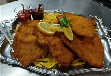 Ein großer Teller mit knusprigem Schnitzel, serviert mit Pommes frites und zwei verschiedenen Dips. Frische Zitronenscheiben und ein Minzblatt verleihen dem Gericht eine dekorative Note.
